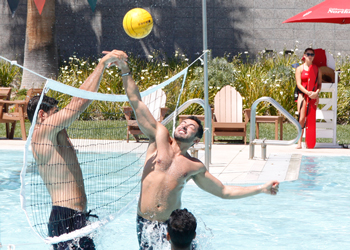 Three male students play water volleyball in the Rec pool while a female lifeguard watches at the Poolside DJ event.